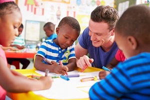 Male nursery practitioner at a table with 3 young children doing a writing activity