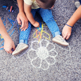 Image of children's legs on the ground and hands using chalk to draw a flower