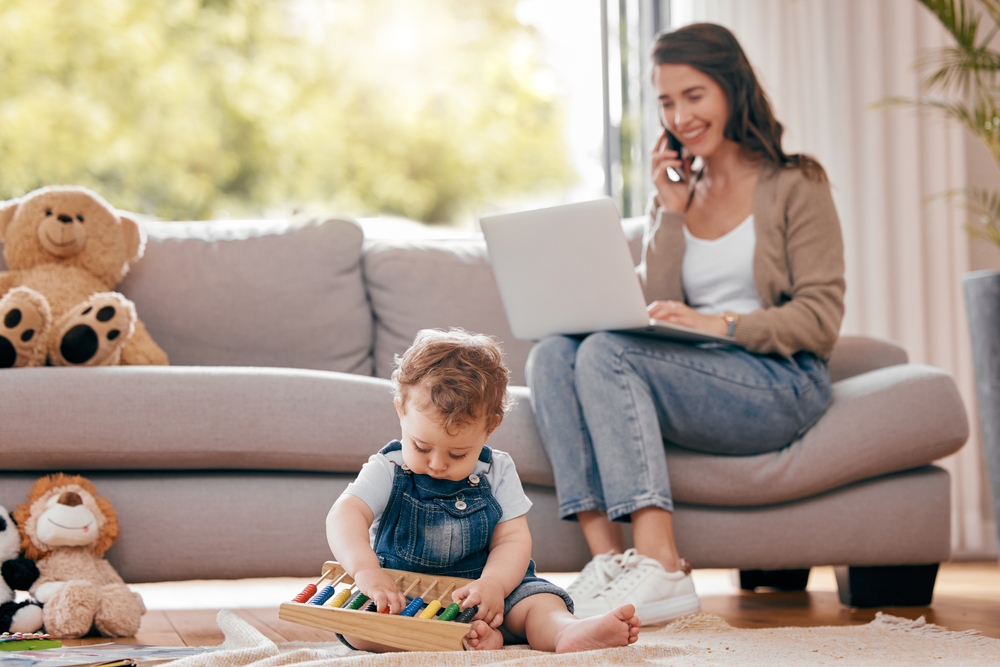 Woman on a phone call with a laptop on her lap watching a toddler play with an abacus