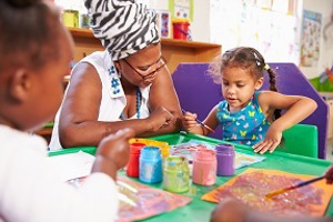 Black female nursery practitioner at a table with 2 young children playing